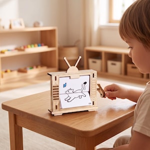 May include: A light-colored wooden mechanical flipbook toy shaped like a vintage television set with an antenna on top. A hand-crank on the side operates the internal mechanism, displaying a paper illustration of a cat and a blue butterfly with the number 09.