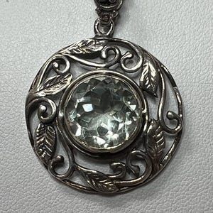 May include: A round silver pendant featuring a central faceted pale green gemstone. The circular frame is decorated with an openwork design of swirling vines and leaves. A decorative silver bail is attached to the top of the circular pendant.