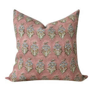 May include: A square throw pillow in a dusty rose pink color featuring a repeating pattern of small, stylized floral motifs in shades of light blue and sage green. The fabric has a visible, slightly textured weave throughout the surface.