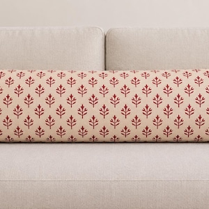 May include: A long, rectangular lumbar pillow featuring a repeating pattern of small, dark red stylized leaf or floral motifs printed across a light beige fabric background. The pillow has a clean, finished edge and a uniform, structured shape.