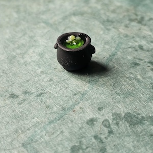 May include: A miniature black cauldron with two small handles, filled with a vibrant green liquid and several small, round, light-colored objects. The cauldron is set against a textured, teal-colored surface, creating a whimsical, fantasy-themed image.