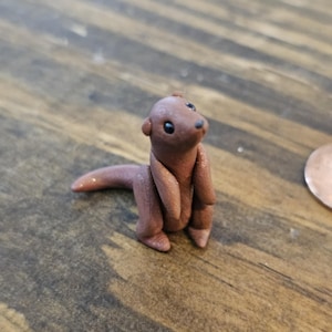 May include: A small, handcrafted brown otter figurine with black eyes and a tail. The otter is posed upright, with its arms and legs detailed. The figurine is set on a wooden surface.