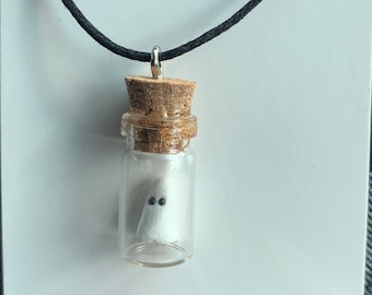 May include: A white ghost with black eyes is inside a small glass bottle with a cork stopper. The bottle is attached to a black cord necklace. The necklace is on a white background with the text "Kitcat's Designs kitcatsdesigns.etsy.com".
