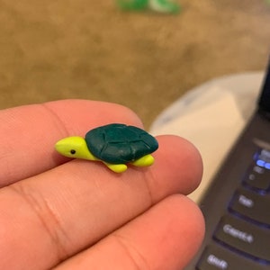 Puede incluir: Pequeña figura de tortuga con caparazón verde oscuro y cabeza y extremidades amarillas. La figura se sostiene en una mano, con un teclado de portátil en el fondo. Adorno pequeño y detallado.