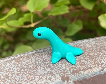May include: A small, turquoise clay sculpture of a Loch Ness Monster, with a long neck, small head, and four flipper-like appendages. The creature has a single black eye and is set against a blurred green background.
