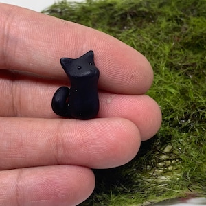May include: A small black cat figurine made of polymer clay. The cat is sitting up with its tail curled around its legs. The figurine is about 1 inch tall.