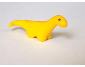 May include: A small, yellow dinosaur figurine with a black eye. The dinosaur is made of a soft, pliable material.