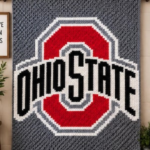 Pode incluir: Uma manta retangular de crochê com fundo cinza e um grande logotipo da Ohio State University centralizado. O logotipo consiste em uma letra O vermelha em bloco contornada em branco e cinza, com o texto preto OHIO STATE no centro.