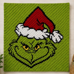 May include: A square crochet blanket featuring a pixelated green character face with yellow eyes and a black outline. The character wears a red Santa hat with a white textured brim and pom-pom, all set against a solid lime green background.