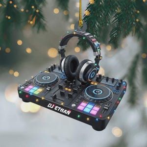 May include: A black DJ controller ornament featuring the text DJ ETHAN, topped with a pair of black and silver headphones. The device is decorated with small, colourful string lights and hangs from a gold cord, creating a festive musical display.