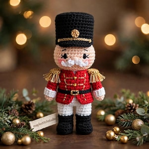May include: A crocheted nutcracker doll featuring a tall black hat with a gold emblem, a red jacket with gold fringe epaulets and buttons, white trousers, and black boots. The doll has a white moustache and a tag reading maplestitchstudio.