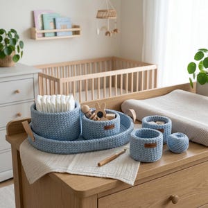 May include: A set of light blue crocheted storage baskets and an oval tray with tan leather-like tabs. The baskets contain white folded diapers and wooden baby toys. A small ball of matching blue yarn and a wooden crochet hook are also present.