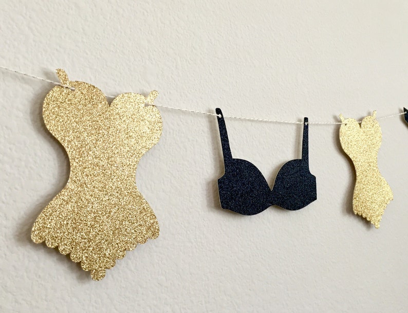 May include: A gold glitter and black glitter paper banner with a corset and bra design. The banner is strung on a white string.