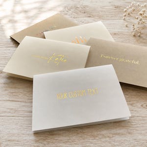 May include: A collection of folded note cards in white, cream, and kraft paper shades. Each card features gold foil-stamped text, including phrases like "Forever grateful," "Katie," and "YOUR CUSTOM TEXT" centered on the front of the cards.