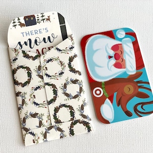 May include: A white envelope with a festive wreath pattern and the text "There's snow place like home" inside. The envelope is open and a gift card featuring Santa Claus and a reindeer is visible.