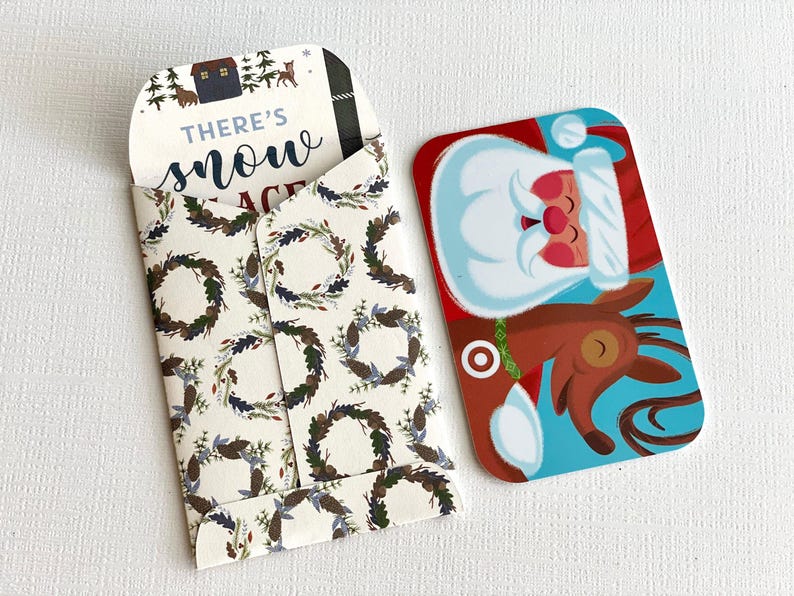 May include: A white envelope with a festive wreath pattern and the text "There's snow place like home" inside. The envelope is open and a gift card featuring Santa Claus and a reindeer is visible.