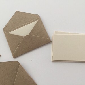 Puede incluir: Un conjunto de sobres peque&ntilde;os de papel texturizado marr&oacute;n claro con solapas triangulares, cada uno con una tarjeta rectangular lisa de color blanco roto. Una pila de tarjetas rectangulares de color blanco roto se sit&uacute;a a la derecha.