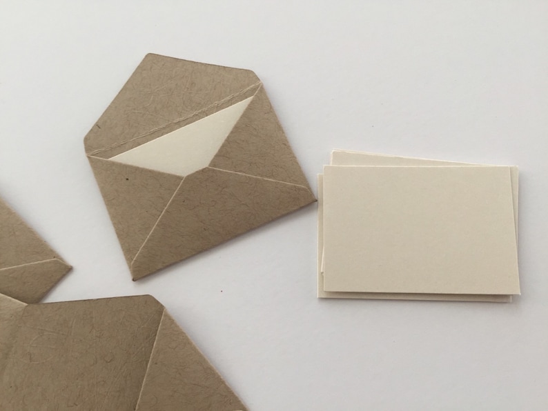 Puede incluir: Un conjunto de sobres peque&ntilde;os de papel texturizado marr&oacute;n claro con solapas triangulares, cada uno con una tarjeta rectangular lisa de color blanco roto. Una pila de tarjetas rectangulares de color blanco roto se sit&uacute;a a la derecha.