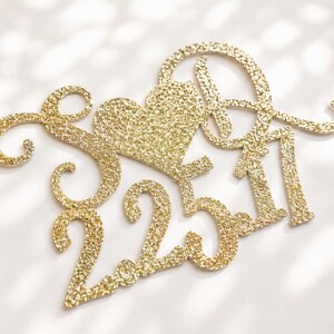 May include: A gold glitter cake topper featuring the letters A and D, a heart shape, and the date 2.25.17. The design includes decorative swirling flourishes on the left side and is constructed from a single piece of textured, sparkling material.