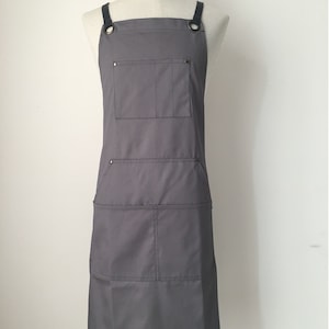 May include: A solid grey apron featuring dark grey shoulder straps attached with metal grommets. The apron includes a single chest pocket and two lower pockets, each divided into two sections. The word men's is printed in black in the top left corner.