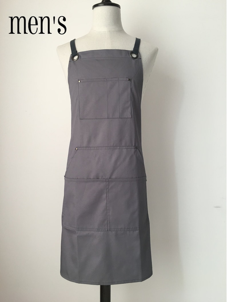 May include: A solid grey apron featuring dark grey shoulder straps attached with metal grommets. The apron includes a single chest pocket and two lower pockets, each divided into two sections. The word men's is printed in black in the top left corner.