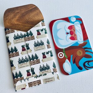 May include: A wooden card holder with a Christmas tree pattern and a Santa Claus gift card. The card holder has a light brown wooden top and a white background with green Christmas trees and the words "North Pole" and "Believe".