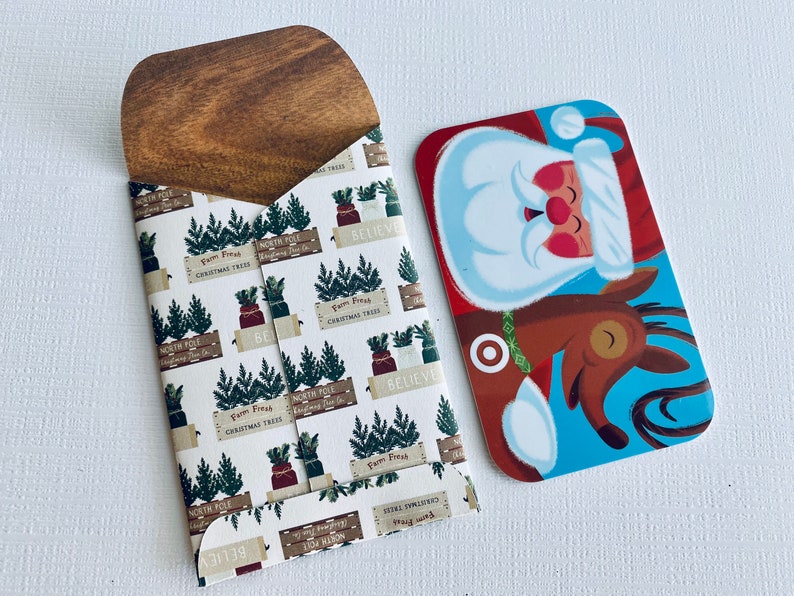 May include: A wooden card holder with a Christmas tree pattern and a Santa Claus gift card. The card holder has a light brown wooden top and a white background with green Christmas trees and the words "North Pole" and "Believe".