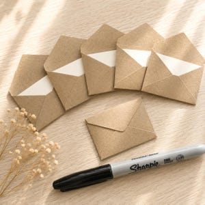 Puede incluir: Cinco sobres abiertos de papel kraft marr&oacute;n con interior blanco dispuestos en abanico, un sobre cerrado de papel kraft marr&oacute;n colocado debajo y un marcador permanente gris Sharpie de punta fina situado horizontalmente.