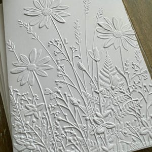 Puede incluir: Una tarjeta de felicitaci&oacute;n blanca con un dise&ntilde;o en relieve de un prado de flores silvestres. La textura representa varias flores, incluyendo margaritas, helechos y hierbas altas, creando un efecto tridimensional en el papel.