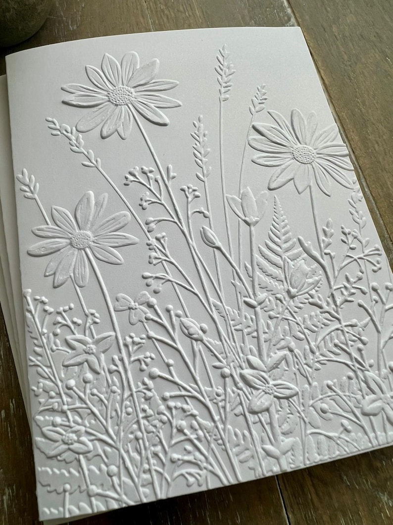 Puede incluir: Una tarjeta de felicitaci&oacute;n blanca con un dise&ntilde;o en relieve de un prado de flores silvestres. La textura representa varias flores, incluyendo margaritas, helechos y hierbas altas, creando un efecto tridimensional en el papel.