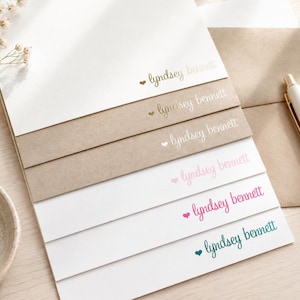 May include: A stack of rectangular stationery cards featuring the printed name lyndsey bennett in various colors and fonts, each accompanied by a small heart icon. The cards are layered on top of each other, displaying white and kraft paper textures.
