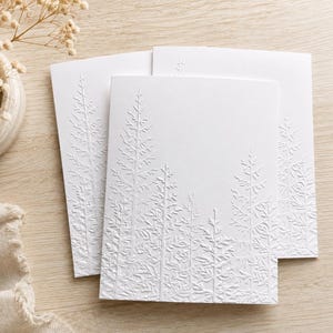 May include: Three white folded greeting cards featuring a subtle, embossed design of evergreen trees on the front cover. The texture of the trees is raised, creating a monochromatic look against the smooth, matte white paper of the card stock.