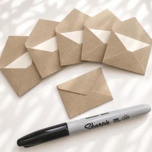 May include: Six small, light brown kraft paper envelopes arranged in a semi-circle with one centered below, accompanied by a grey Sharpie fine point permanent marker with a black cap, positioned horizontally at the bottom of the frame.