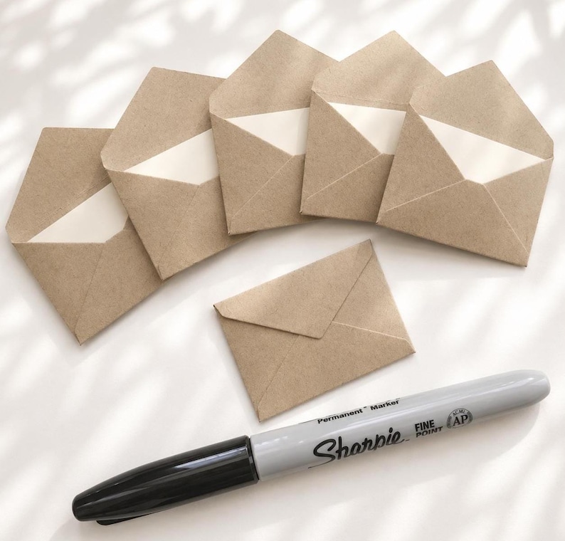 Puede incluir: Seis sobres peque&ntilde;os de papel kraft marr&oacute;n claro dispuestos en semic&iacute;rculo con uno centrado debajo, acompa&ntilde;ados por un marcador permanente Sharpie gris de punta fina con tapa negra, colocado horizontalmente en la parte inferior.