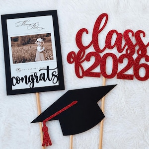 May include: A graduation-themed photo frame, a red glitter "Class of 2026" topper, and a black mortarboard with a red tassel. The photo frame has a black border with a photo and the word "congrats". All items are on wooden sticks.