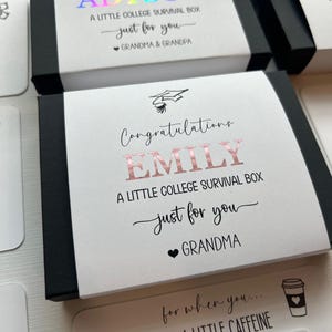 Op de afbeelding: Een zwarte rechthoekige geschenkdoos met een witte papieren hoes met de tekst Congratulations EMILY A LITTLE COLLEGE SURVIVAL BOX just for you GRANDMA met een afstudeerhoed-icoon en een hartje in ros&eacute;gouden foliebedrukking.