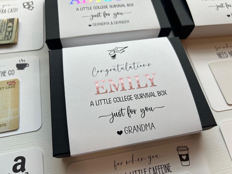 Op de afbeelding: Een zwarte rechthoekige geschenkdoos met een witte papieren hoes met de tekst Congratulations EMILY A LITTLE COLLEGE SURVIVAL BOX just for you GRANDMA met een afstudeerhoed-icoon en een hartje in ros&eacute;gouden foliebedrukking.