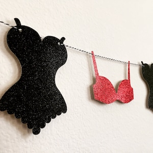 May include: A black and red glitter garland with a black corset, red bra, and black lingerie cutouts. The garland is strung on a white string with black and white twine.