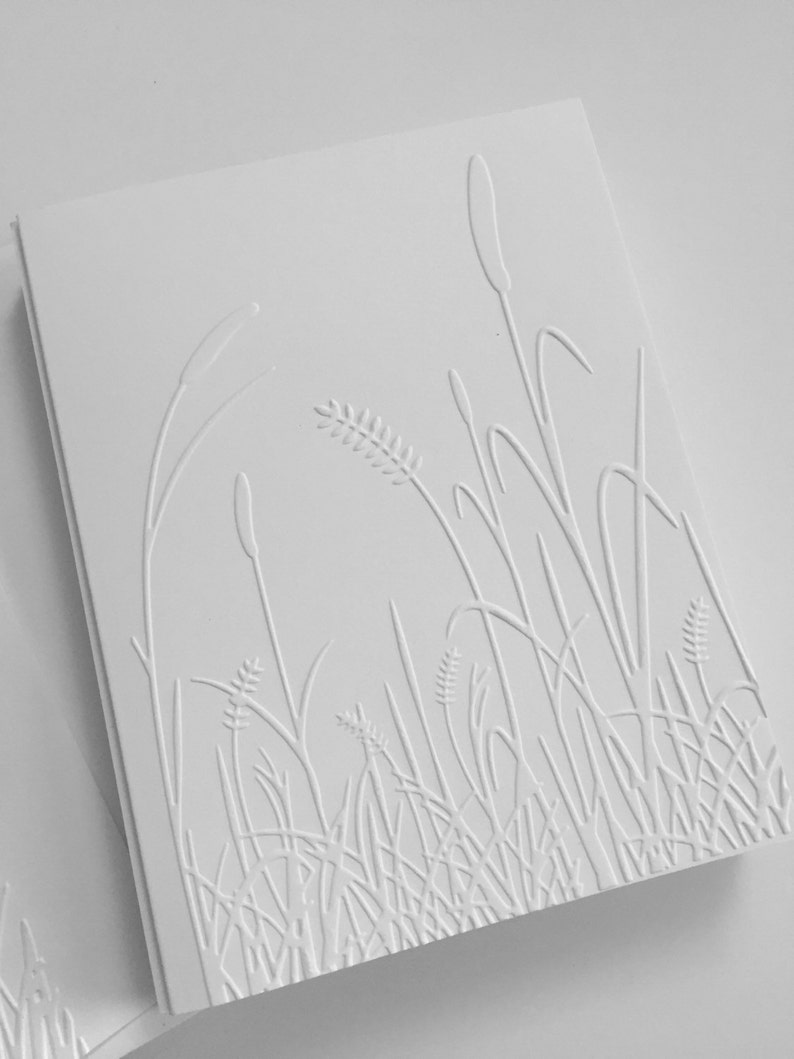 Op de afbeelding: Een witte wenskaart met een verhoogd, reli&euml;fontwerp van hoog gras en riet. De textuur is gecre&euml;erd door middel van een blinddruktechniek, wat resulteert in een monochroom, tactiel oppervlak dat de botanische vormen benadrukt.