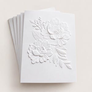 May include: A stack of white greeting cards featuring a raised, embossed floral design on the front cover. The embossed pattern displays two large, detailed flowers surrounded by leaves and small sprigs, creating a textured, monochromatic look.
