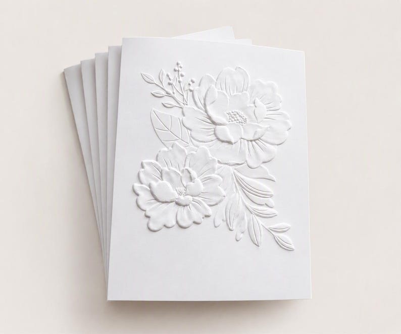 May include: A stack of white greeting cards featuring a raised, embossed floral design on the front cover. The embossed pattern displays two large, detailed flowers surrounded by leaves and small sprigs, creating a textured, monochromatic look.