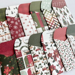 May include: A collection of Christmas-themed paper envelopes with various patterns. Designs include reindeer, Christmas trees, snowflakes, and floral motifs in red, green, and white. Some envelopes have text that reads "Merry everything & happy always."