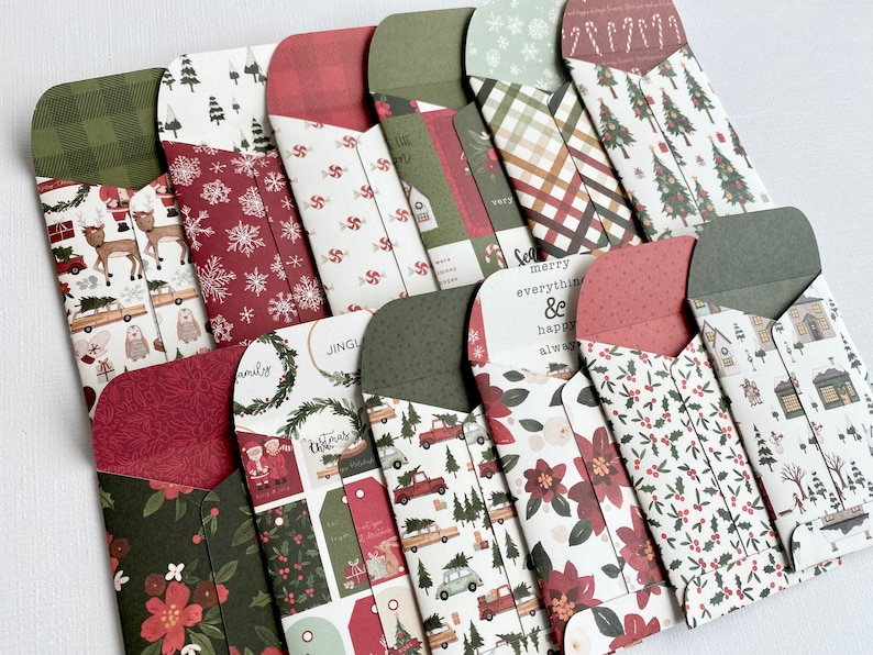 May include: A selection of Christmas-themed paper envelopes with diverse patterns. Designs include reindeer, Christmas trees, snowflakes, and floral motifs in red, green, and white. Some envelopes have text that reads "Merry everything & happy always."