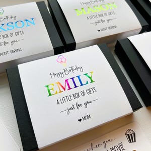 May include: Several black rectangular gift boxes wrapped in white paper sleeves. Each sleeve features a colorful foil-stamped name, the text "Happy Birthday," "A LITTLE BOX OF GIFTS," "just for you," and a sender name with a heart icon.