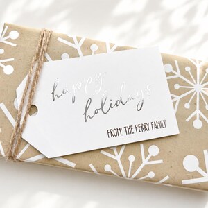 May include: A rectangular gift wrapped in brown paper with a white snowflake pattern, tied with brown twine. A white rectangular gift tag is attached, featuring the silver foil text "happy holidays" and the black printed text "FROM: THE PERRY FAMILY".