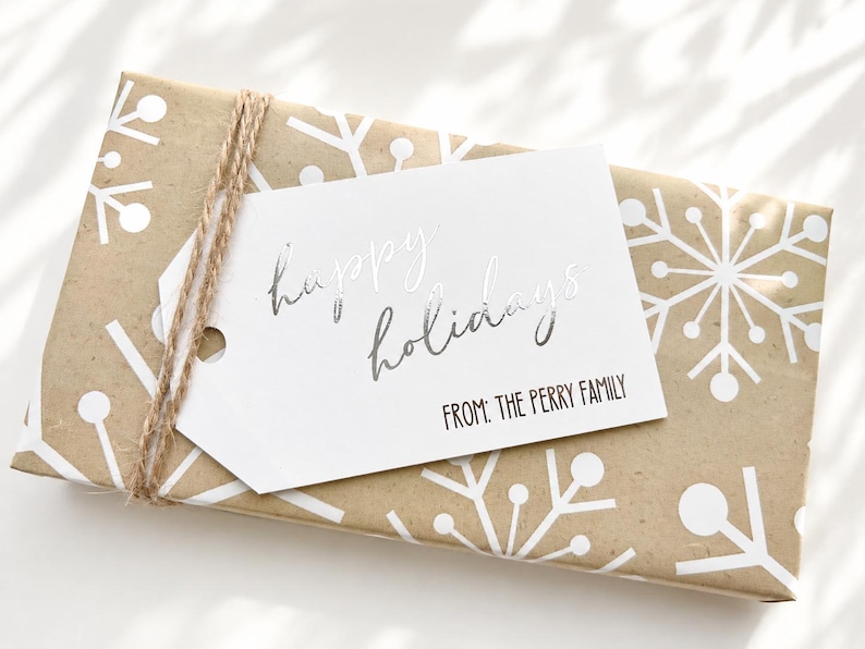 May include: A rectangular gift wrapped in brown paper with a white snowflake pattern, tied with brown twine. A white rectangular gift tag is attached, featuring the silver foil text "happy holidays" and the black printed text "FROM: THE PERRY FAMILY".