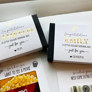 Op de afbeelding: Twee rechthoekige zwarte doosjes met witte hoezen voorzien van goudfolie tekst die luidt Congratulations CAMERON A LITTLE COLLEGE SURVIVAL BOX just for you MOM en Congratulations EMILY A LITTLE COLLEGE SURVIVAL BOX just for you GRANDMA.