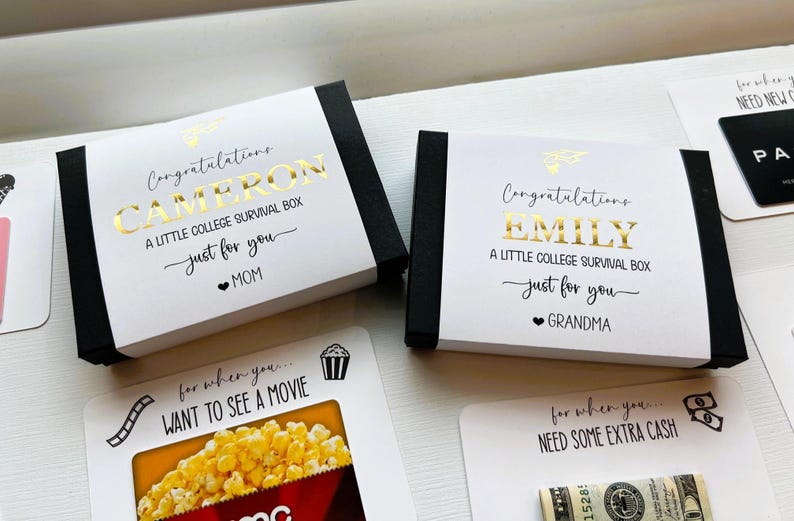 Op de afbeelding: Twee rechthoekige zwarte doosjes met witte hoezen voorzien van goudfolie tekst die luidt Congratulations CAMERON A LITTLE COLLEGE SURVIVAL BOX just for you MOM en Congratulations EMILY A LITTLE COLLEGE SURVIVAL BOX just for you GRANDMA.