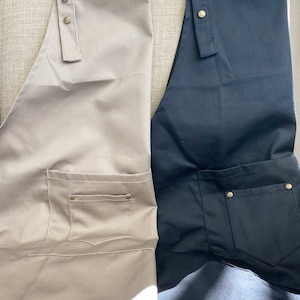 May include: Two aprons, one in beige and one in black, each featuring a front pocket with metallic rivets and adjustable shoulder straps secured by three brass-colored metal snaps at the top. The fabric has a smooth, uniform texture.