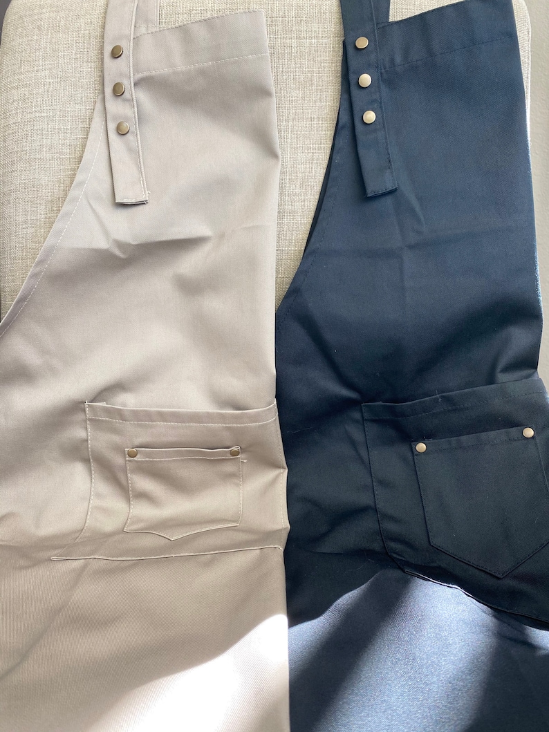 May include: Two aprons, one in beige and one in black, each featuring a front pocket with metallic rivets and adjustable shoulder straps secured by three brass-colored metal snaps at the top. The fabric has a smooth, uniform texture.